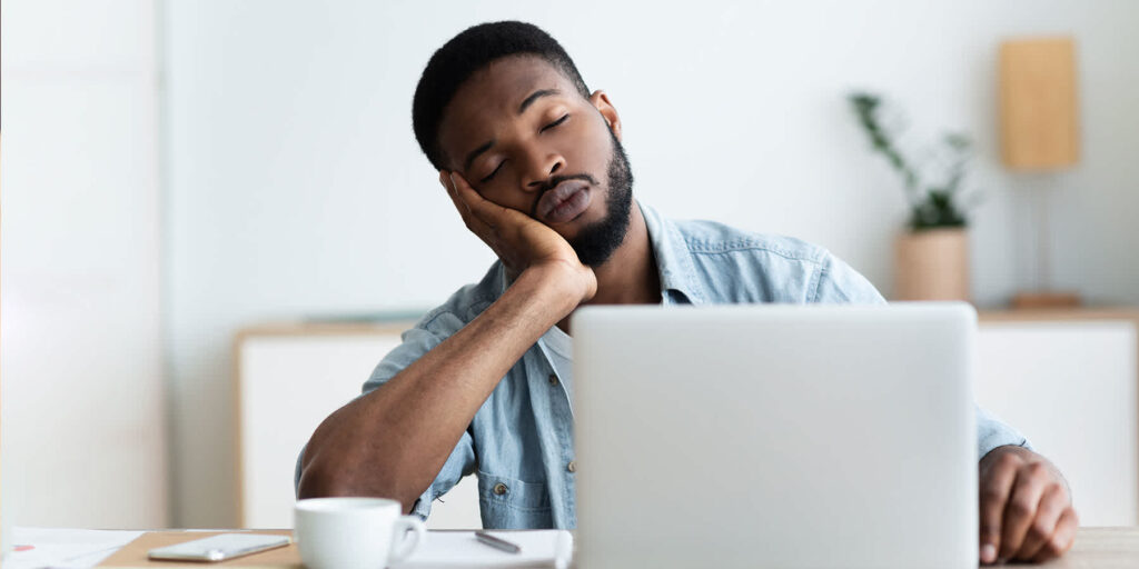 Tired young man sleeping at his desk with laptop, showing signs of fatigue and potential nutrient deficiency in a home office setting."