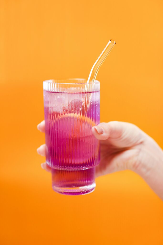 Vibrant purple drink in a glass with a straw held against orange background.